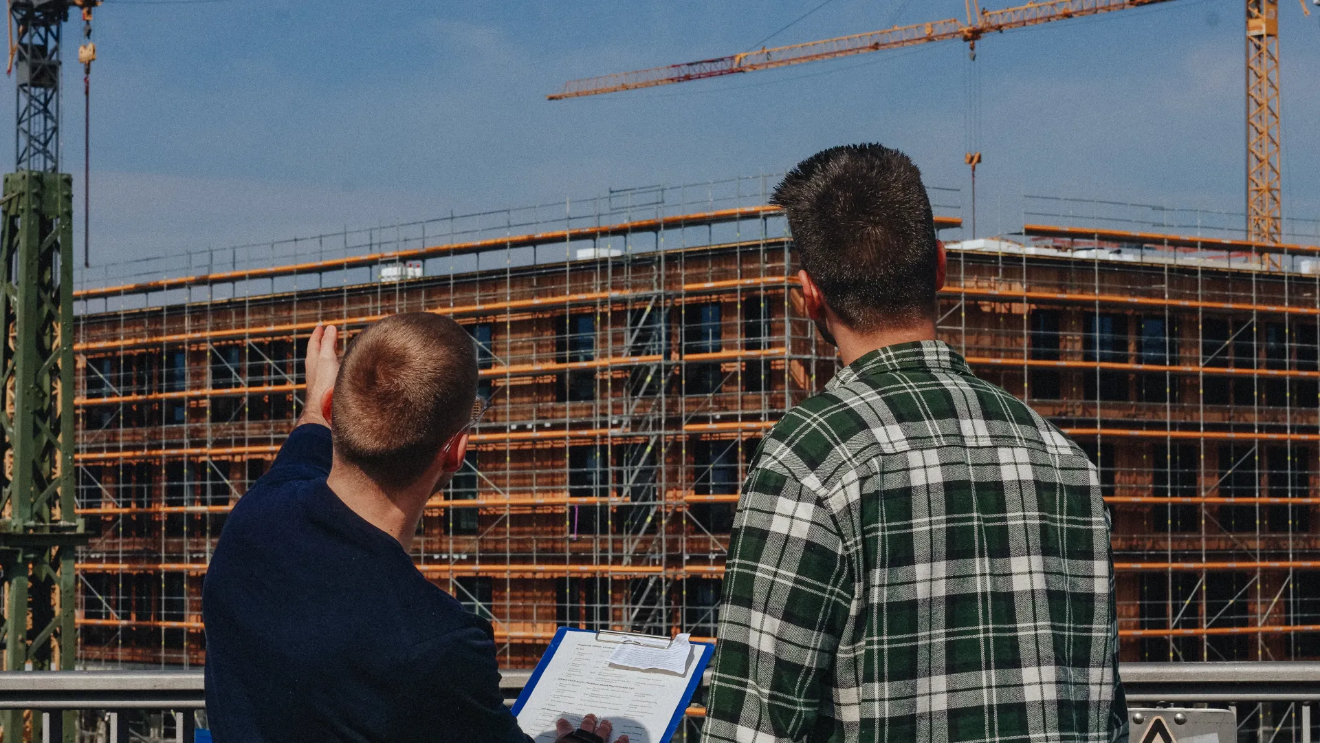 Zwei Personen betrachten eine Baustelle mit Rohbau und Kränen im Hintergrund.